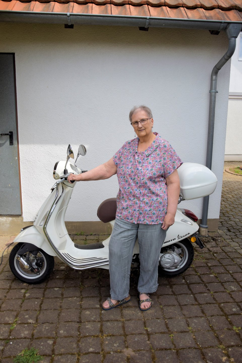Frau mit Bluse aus Abby und grauer Hose vor weißem Motorroller